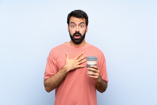 Jeune homme à la barbe tenant un café à emporter sur bleu isolé surpris et choqué tout en regardant à droite