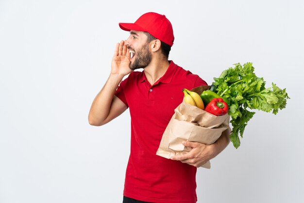 Jeune homme à la barbe tenant un sac plein de légumes isolé sur blanc en criant avec la bouche grande ouverte