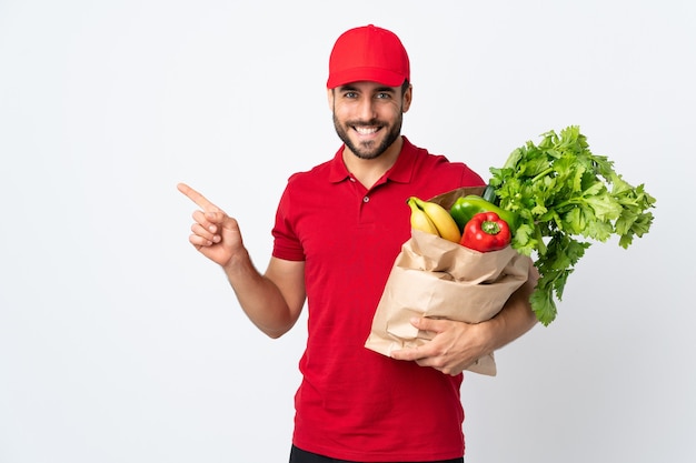 Jeune homme à la barbe tenant un sac plein de légumes isolé sur blanc doigt pointé sur le côté