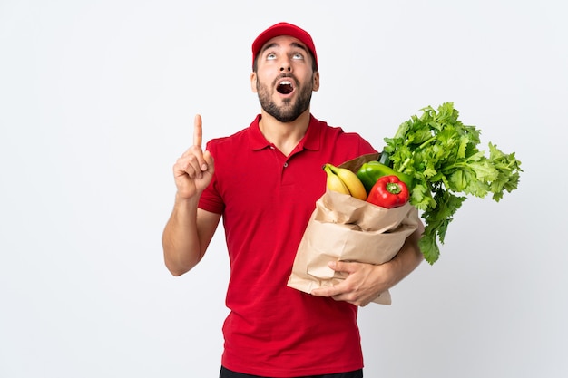 Jeune homme à la barbe tenant un sac plein de légumes isolé sur un mur blanc pointant avec l'index une excellente idée