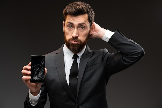 Jeune homme à la barbe tenant un smartphone cassé avec une expression confuse sur son visage et pensant quoi faire. Stock photo