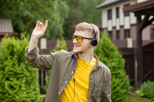 Jeune homme barbu accueille quelqu'un avec vague de sa main, écouter de la musique en ligne grâce à des écouteurs modernes à l'extérieur en plein air sur la journée d'été ensoleillée