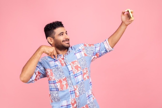 Jeune homme barbu adulte en chemise bleue parlant par appel vidéo et pointant vers le bas pour s'abonner ayant une conversation en ligne sur téléphone mobile prenant selfie Prise de vue en studio intérieur isolée sur fond rose