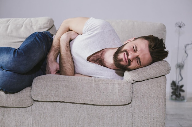 Jeune homme barbu allongé sur un canapé tenant son ventre à cause d'une douleur intense