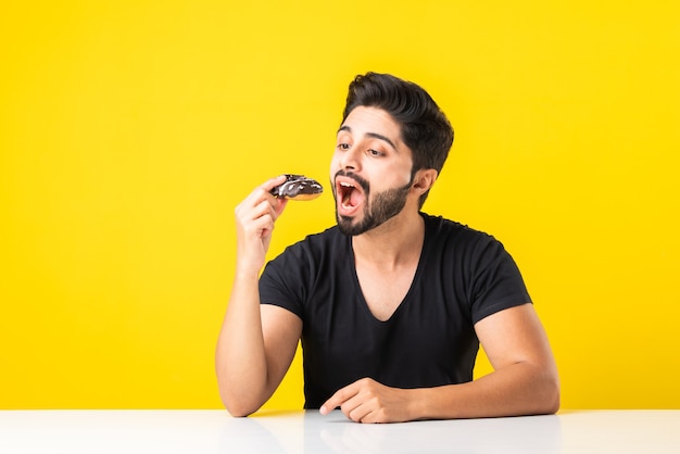 Jeune homme barbu asiatique indien mange un beignet ou un beignet assis à la table de la cuisine sur fond jaune ou blanc