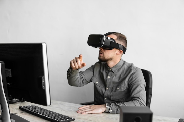 Jeune homme barbu assis contre un ordinateur avec des lunettes de réalité virtuelle essayant de toucher quelque chose