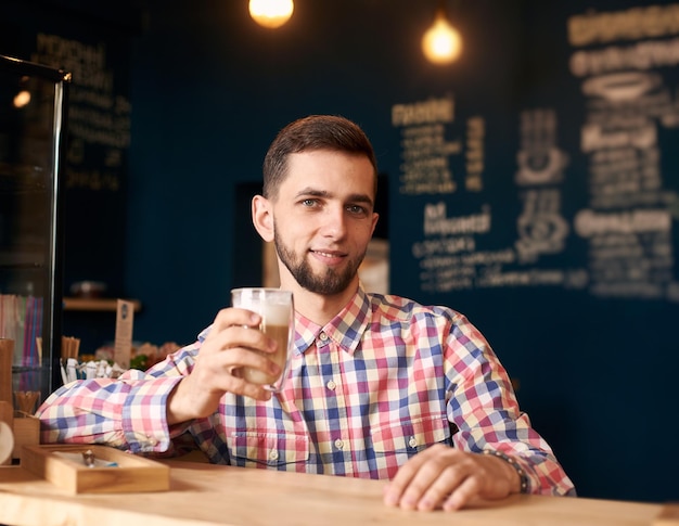 Jeune homme barbu assis dans un café et souriant à huis clos tenant une tasse de café au lait Prise de vue en portrait Fond sombre