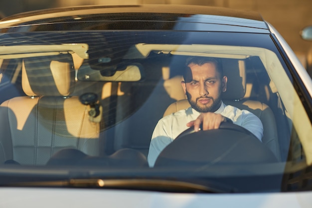 Jeune homme barbu assis dans sa voiture et regardant la caméra pendant la conduite