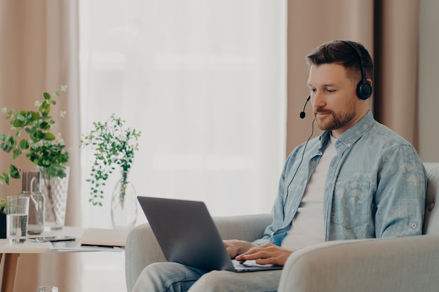 Jeune homme barbu attentif dans un casque tapant sur un ordinateur portable pendant une conférence en ligne alors qu'il était assis dans un fauteuil à la maison, un pigiste travaillant à distance. Concept de travail à distance