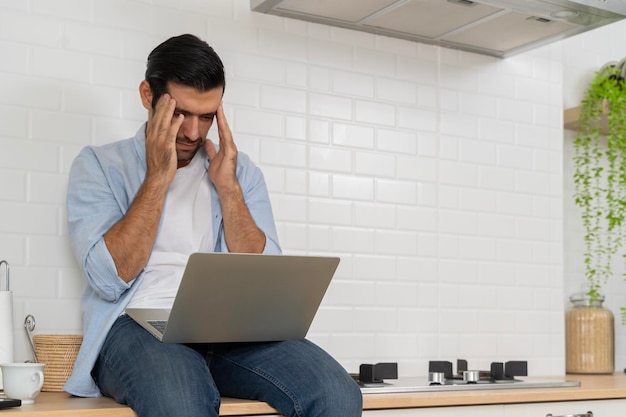 Un jeune homme barbu attrayant utilise un ordinateur portable pour surfer sur les réseaux sociaux ou vérifier son travail tout en étant assis sur le comptoir de la cuisine à la maison avec un espace de copie stressant