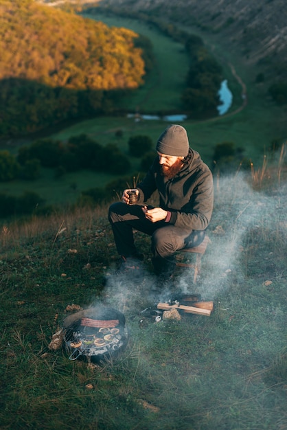 Jeune homme barbu sur une colline est assis près d'un grill avec un café