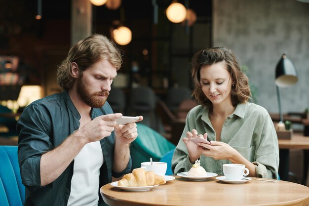 Jeune homme barbu concentré en chemise décontractée buvant du café et mangeant un croissant tout en vérifiant s