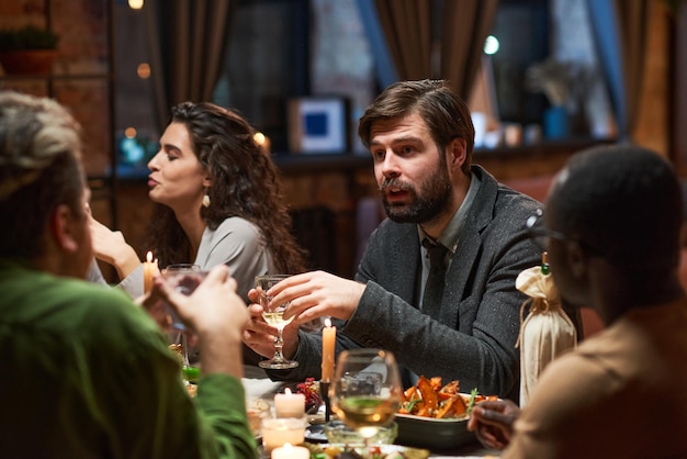 Jeune homme barbu en costume de parler aux gens alors qu'il était assis à table à manger pendant le dîner