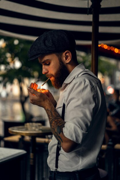 Jeune homme barbu dans un café dans la rue fume une cigarette. Mec romantique dans une casquette de chemise blanche et des bretelles dans la ville. Peaky Blinders. vieux rétro à la mode.