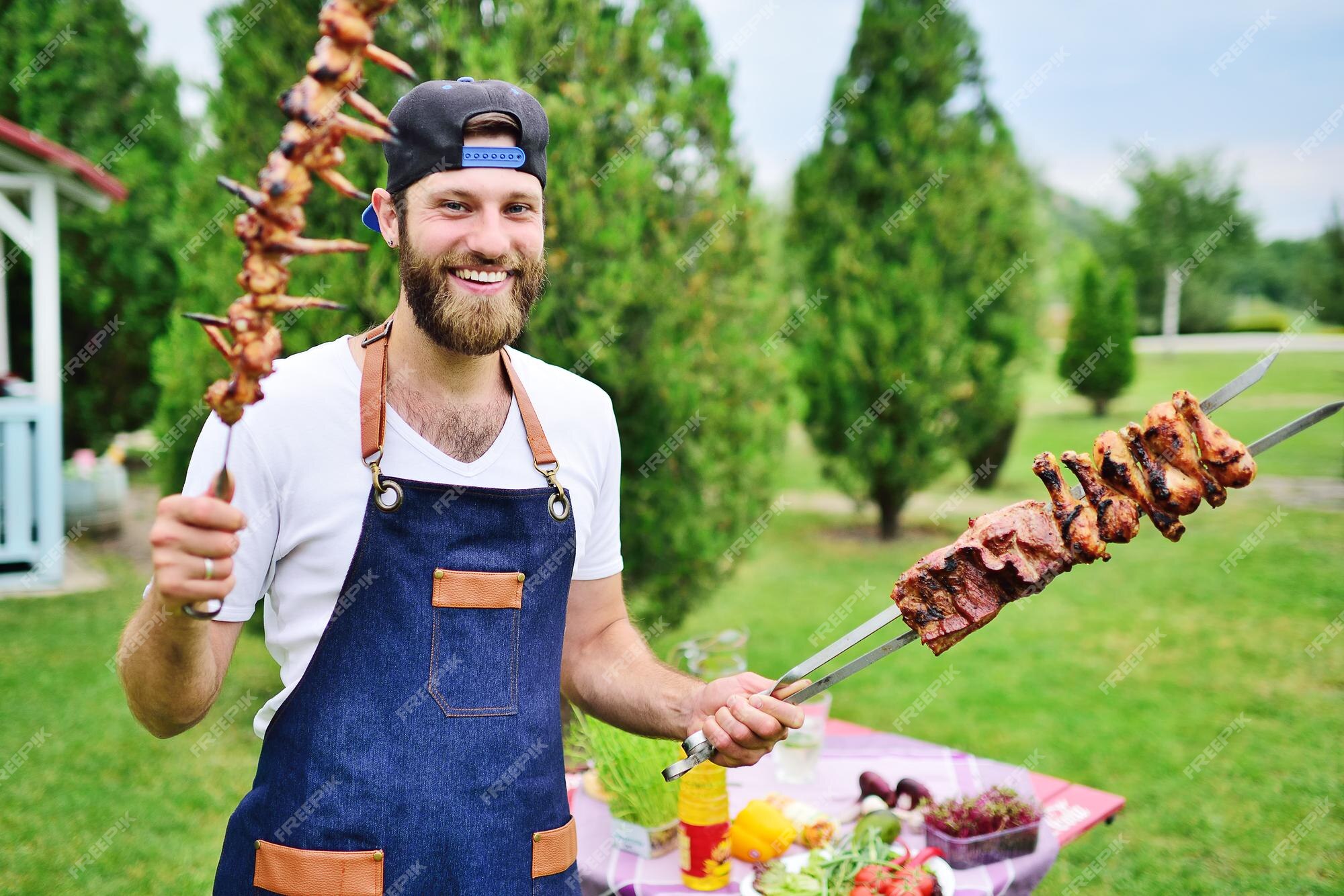 Jeune Homme Barbu Dans Une Casquette Et Un Tablier En Denim Sourit Et Tient  Une Brochette Avec De Délicieuses Viandes De Barbecue Co