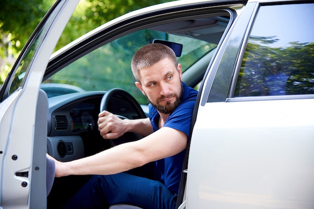 Un jeune homme barbu est assis dans une voiture avec une porte ouverte et regarde en arrière.