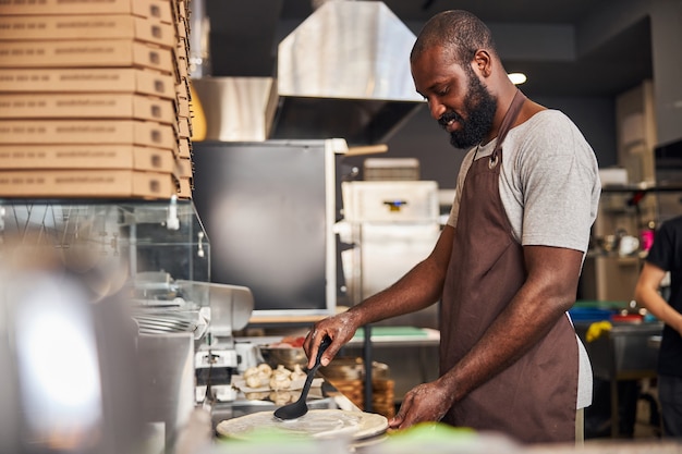 Un jeune homme barbu est debout à table et répand de la sauce sur de la pâte avec une cuillère dans la cuisine