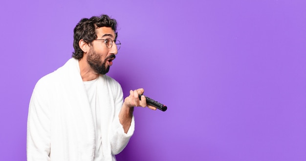 Jeune homme barbu fou portant un peignoir avec une télécommande de télévision