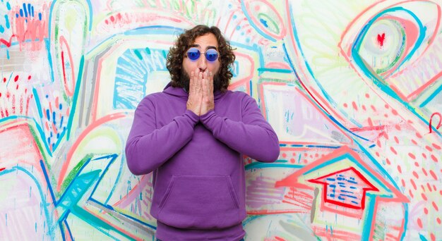 Jeune homme barbu fou se sentant inquiet, bouleversé et effrayé, se couvrant la bouche avec les mains, semblant anxieux et s'être égaré contre un mur de graffitis