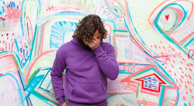 Jeune homme barbu fou se sentant triste, frustré, nerveux et déprimé, couvrant le visage à deux mains, pleurant contre un mur de graffitis