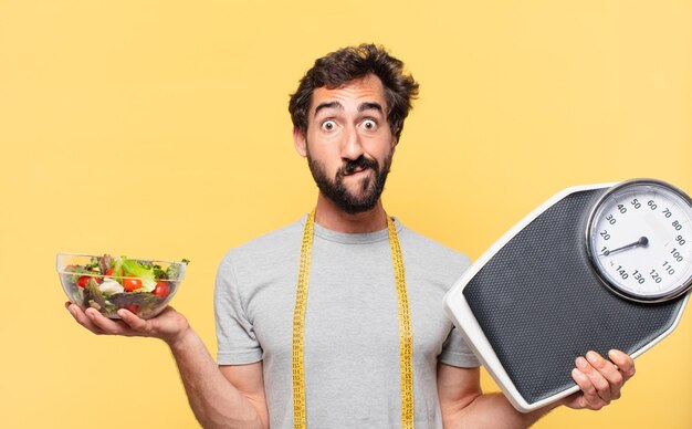 Jeune Homme Barbu Fou Suivant Un Régime Doutant Ou Incertain D'expression Et Tenant Une Balance Et Une Salade