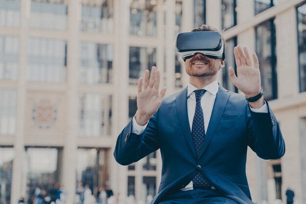 Jeune homme barbu heureux employé de bureau habillé formellement assis à l'extérieur et utilisant un casque VR ou des lunettes 3d