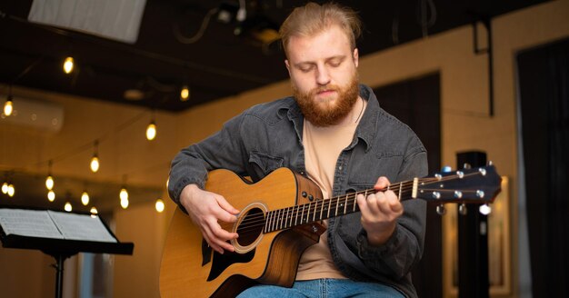 Un jeune homme barbu joue de la guitare acoustique dans une pièce à l'éclairage chaleureux