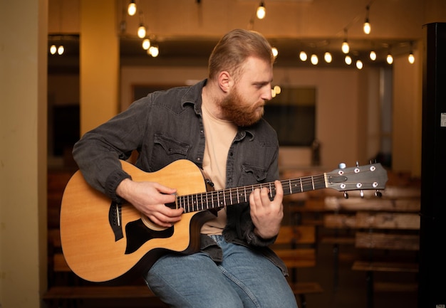 Un jeune homme barbu joue de la guitare acoustique dans une pièce à l'éclairage chaleureux