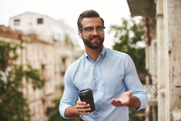 Jeune homme barbu à lunettes et écouteurs tenant une tasse de café et parlant avec un ami