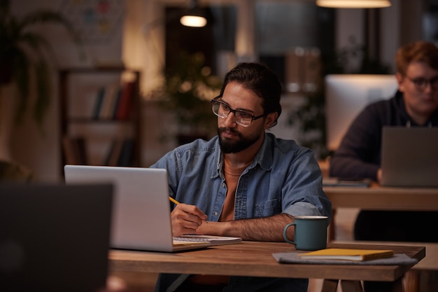 Jeune homme barbu à lunettes, prendre des notes et travailler sur un ordinateur portable à la table au bureau