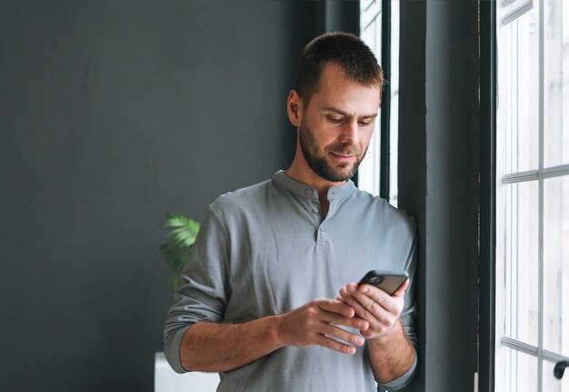 Jeune homme barbu en manches longues grises décontractées avec smartphone dans les mains lisant un message debout à la fenêtre dans un bureau moderne aux murs sombres