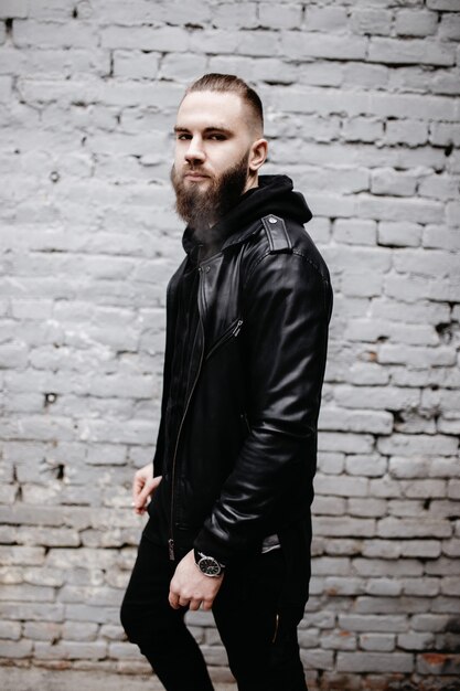 Jeune homme barbu moderne dans des vêtements de style noir posant contre le mur de briques.