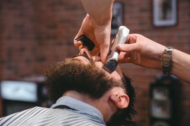 Jeune homme barbu pendant le toilettage de la barbe à l'aide d'une tondeuse dans un salon de coiffure