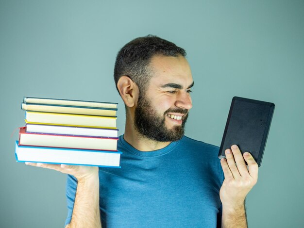 jeune homme barbu avec une pile de livres dans une main et un livre électronique dans l'autre