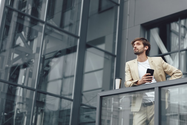 Jeune homme barbu portant un costume à la mode, debout sur le balcon de son propre bureau et à l'aide de smartphone