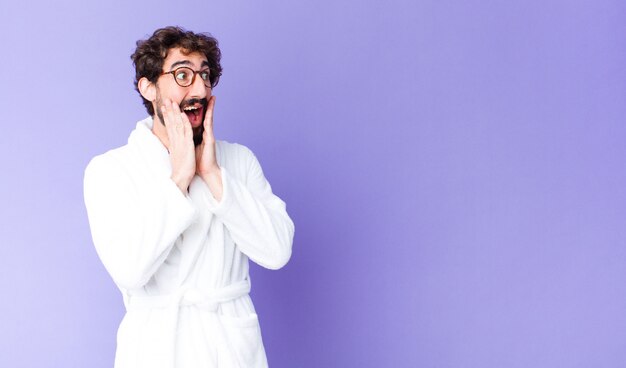 Jeune homme barbu portant un peignoir, heureux, excité et surpris, regardant sur le côté avec les deux mains sur le visage