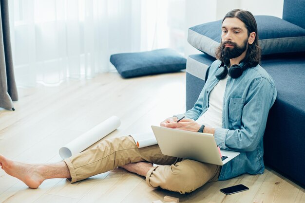 Jeune homme barbu regardant pensivement au loin tout en étant assis sur le sol dans des vêtements décontractés avec un ordinateur portable