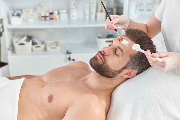 Un jeune homme barbu se détend pendant que le cosmétologue répand de l'argile blanche sur son visage avec une brosse au salon de beauté.