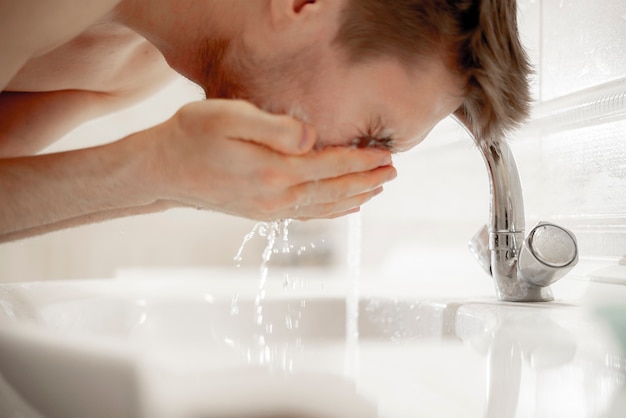 Un jeune homme barbu se lave le visage avec des éclaboussures dans le lavabo de la salle de bain, routine matinale quotidienne