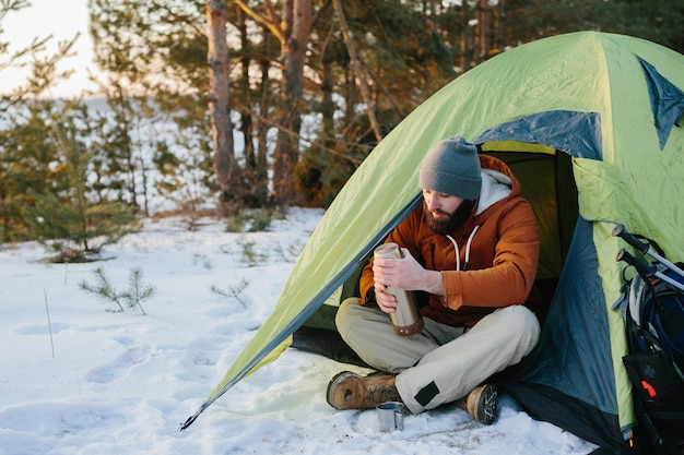 Un jeune homme barbu se repose dans les montagnes hivernales près d'une tente Un voyageur avec une barbe dans une casquette et une veste chaude se réchauffe en buvant du thé ou du café chaud après une randonnée