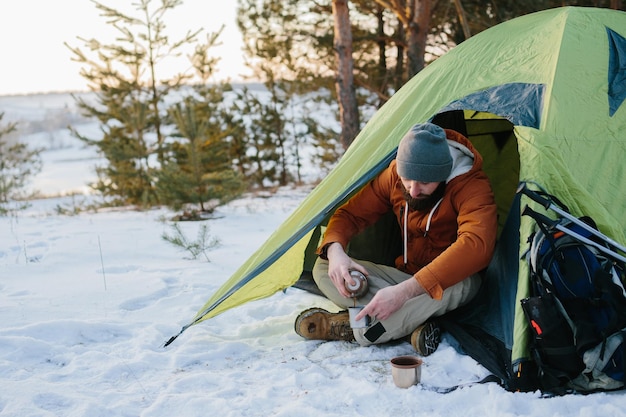 Un jeune homme barbu se repose dans les montagnes hivernales près d'une tente Un voyageur avec une barbe dans une casquette et une veste chaude se réchauffe en buvant du thé ou du café chaud après une randonnée