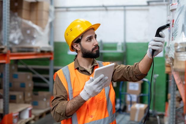 Jeune homme barbu sérieux dans un casque et des gants scannant le code de l'article tout en faisant l'inventaire des stocks dans le magasin