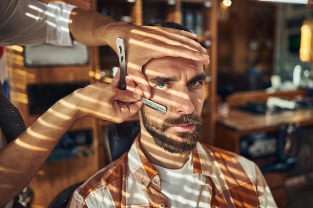 Jeune homme barbu sévère ayant un barbier raser sa barbe avec un rasoir droit dans un salon de coiffure