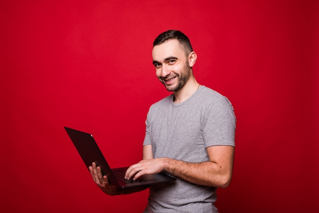 Jeune homme barbu souriant tout en tenant et en utilisant un ordinateur portable isolé sur fond rouge