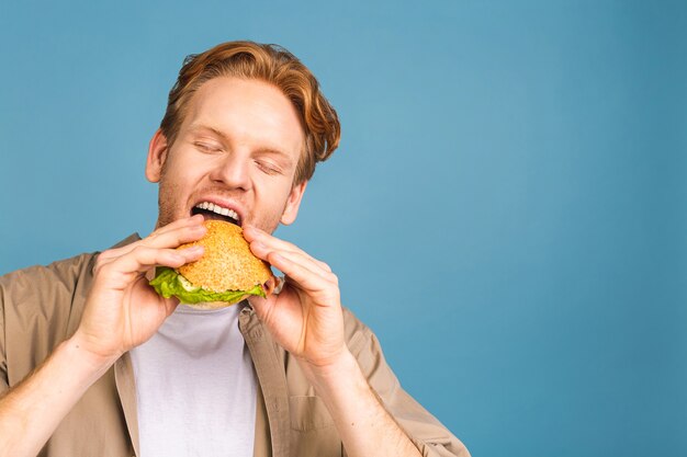 jeune homme barbu tenant un morceau de hamburger. un étudiant mange de la restauration rapide