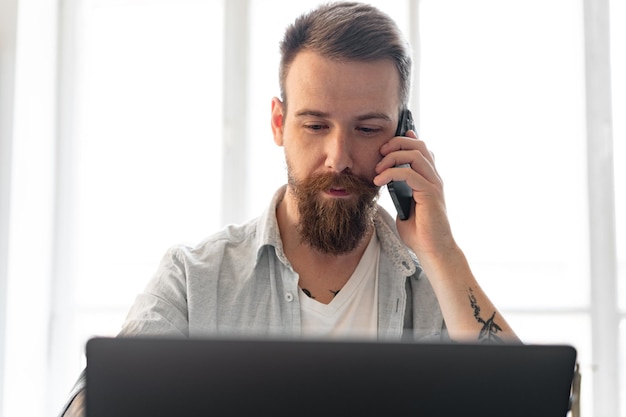 Jeune homme barbu travaillant à domicile avec un ordinateur portable tout en parlant au téléphone