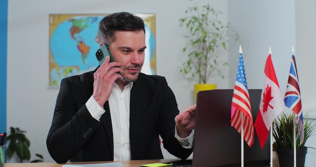 Jeune homme barbu travaille dans une agence de voyage parle au téléphone et regarde un écran d'ordinateur assis à une table sur le fond d'une carte du monde