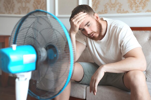 Jeune homme barbu utilisant un ventilateur électrique à la maison assis sur un canapé pour se rafraîchir par temps chaud