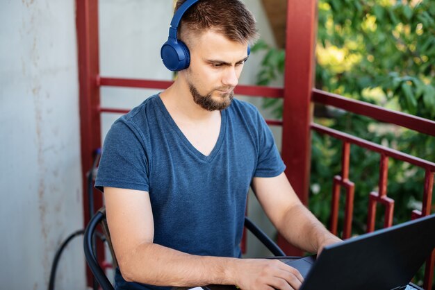 Un jeune homme barbu utilise un ordinateur portable et des écouteurs pour travailler à l'extérieur, travaille sur un projet indépendant en ligne à l'extérieur à la maison, un bel étudiant étudie, apprend une langue, éducation en ligne