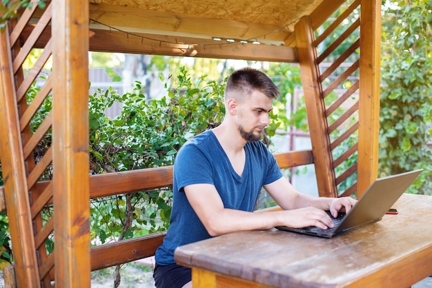 Un jeune homme barbu utilise un ordinateur portable pour travailler à l'extérieur, travaille un projet indépendant en ligne à l'extérieur de la maison, un bel étudiant étudie, apprend une langue, un concept d'éducation en ligne